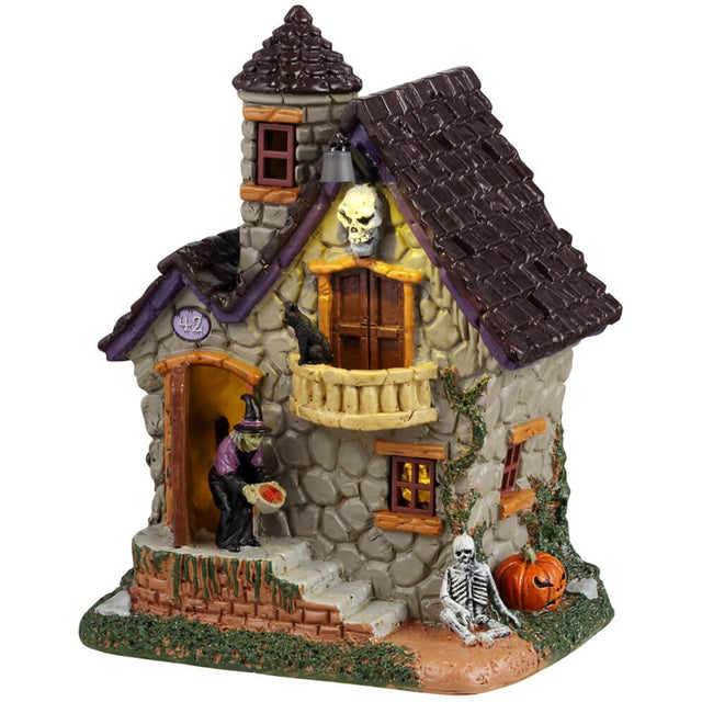 Resin building for a Lemax display. Designed to look like a stone house with moss growing on the side. There's a witch handing out candy at the front door and a skeleton sitting on the ground next to a pumpkin.
