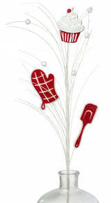 This image shows the pick spray sticking out of a glass vase. The spray is decorated with white balls, white branches, a white and red cupcake, a red and white oven mitt and a red and white spatula.