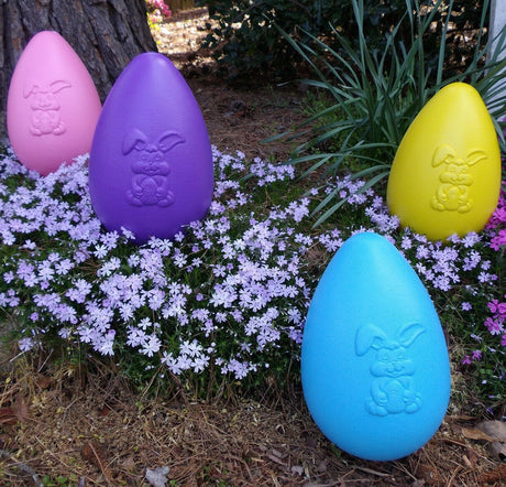 Four blow mold Easter egg decorations in a garden up against a tree. The eggs are pink, purple, blue and yellow and feature a smiling bunny