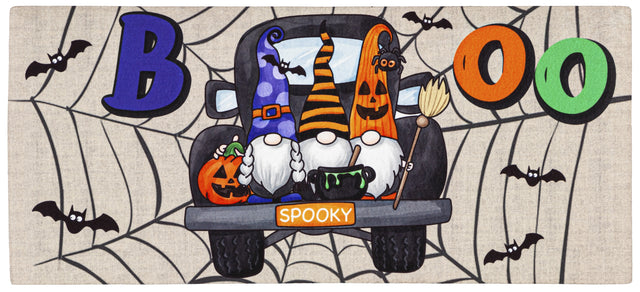 Doormat for Halloween. Reads "BOO" and features three gnomes in the back of a truck. One is holding a pumpkin, one is holding a cauldron and one is holding a broomstick. The license plate reads "SPOOKY" and there are bats and spiderweb accents.