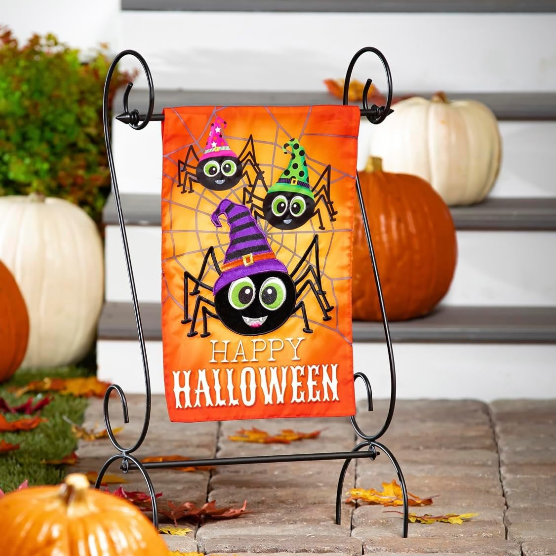 Outdoor Halloween flag. The background is orange and has a gray spiderweb. There are three large, smiling spiders and a message that reads "HAPPY HALLOWEEN." The spiders are all wearing colorful hats. One is pink, one is lime green and one is purple.