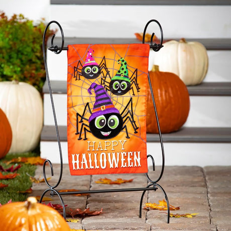 Outdoor Halloween flag. The background is orange and has a gray spiderweb. There are three large, smiling spiders and a message that reads "HAPPY HALLOWEEN." The spiders are all wearing colorful hats. One is pink, one is lime green and one is purple.