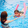 This is a lifestyle image showing a young girl learning to swim using a pink noodle while her mom helps.