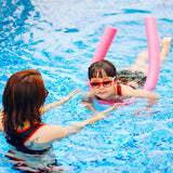 This is a lifestyle image showing a young girl learning to swim using a pink noodle while her mom helps.