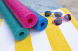 This is a lifestyle image showing three pool noodles laid out on a yellow and white striped beach towel. The pool noodles are teal, pink and blue. On the towel, there are also two pairs of sunglasses: one is blue and one is pink.