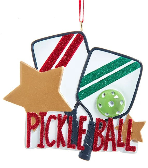 Ornament shaped like two white paddles with festive red and green stripes, as well as two gold stars. It reads "Pickleball" in red glitter.