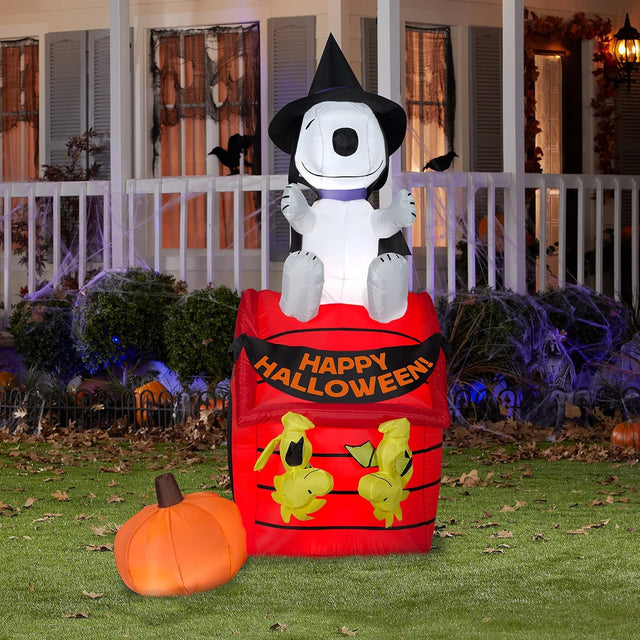 Halloween inflatable shaped like Snoopy the Peanuts cartoon character dressed as a witch and sitting on his red dog house. There are two Woodstock Peanuts cartoon characters hanging from the house like they're bats and there's a pumpkin off to the side.