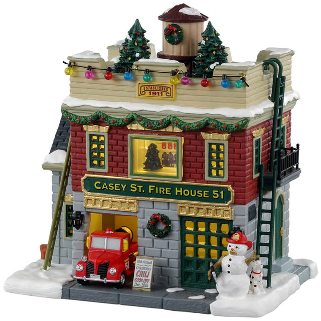 Porcelain lighted building for a Lemax display. Designed to look like a fire house decorated for Christmas. There are decorated trees and multi-colored lights on the roof. There are ladders on both sides of the building. In the window, you can see a decorated tree inside. There's a red firetruck parked in the garage and a snowman on the sidewalk.
