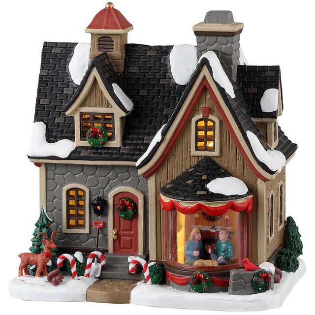 Porcelain lighted building for a Lemax display. Designed to look like a cute home decorated for Christmas. There's snow on the roof and on the sidewalk. There are wreaths, red ribbons and candy cane accents. There's a reindeer and a squirrel near the front door. In the window, you can see a couple relaxing near a fireplace.