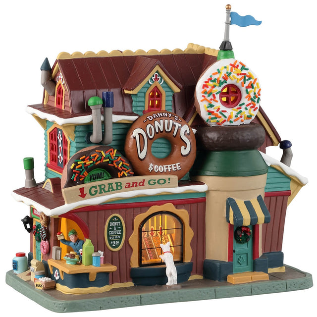 Porcelain lighted building for a Lemax display. Designed to look like a donut shop. There are multiple, large donut statues in front. There's a dog looking in the window at all the treats inside. There's also a vendor behind the booth waving to get your attention.