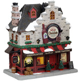 Porcelain lighted building for a Lemax display. Designed to look like a wine and chocolate tasting restaurant. There's a statue of a wine bottle, a wine glass and a piece of chocolate on the front. There are two decorated trees near the front door and a bunch of barrels on the sidewalk.