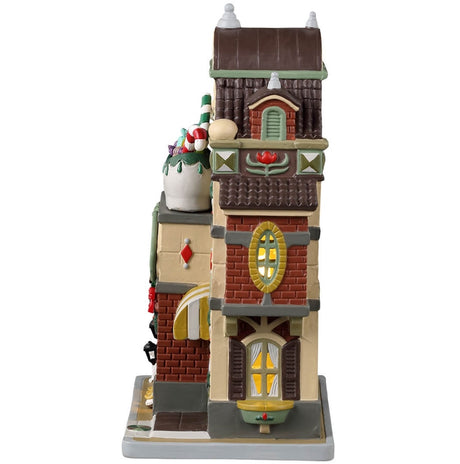 Porcelain lighted building for a Lemax display. Designed to look like a brick shoe store. There's a large wooden shoe statue with gifts inside on the roof. Santa is near the front, offering to help you find the perfect shoes. There's also a white dog on the sidewalk.