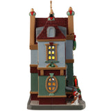 Porcelain lighted building for a Lemax display. Designed to look like a sweater shop. There are a string of sweaters hanging in front and a display of sweaters on the sidewalk. In the window you can see people trying on sweaters. There's a dog wearing a Santa hat waiting at the front door.