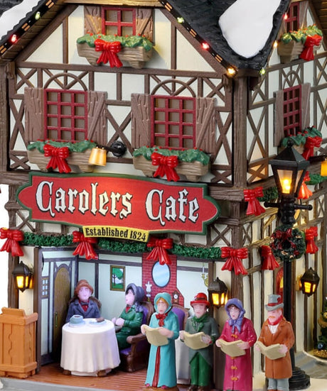 Resin lighted building for a Lemax display. Designed to look like a tall, old-timey cottage decorated for Christmas. There's snow and a string of lights on the roof. There are carolers singing to people dining outside. There are garlands, red ribbons and a decorated Christmas tree.