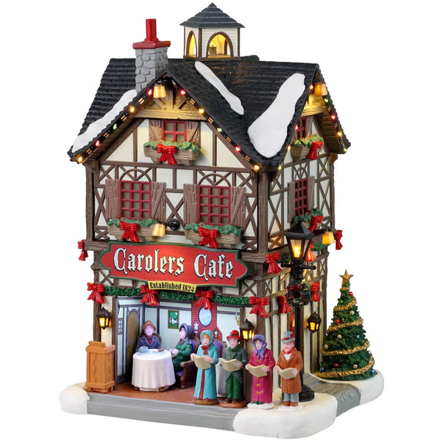 Resin lighted building for a Lemax display. Designed to look like a tall, old-timey cottage decorated for Christmas. There's snow and a string of lights on the roof. There are carolers singing to people dining outside. There are garlands, red ribbons and a decorated Christmas tree.