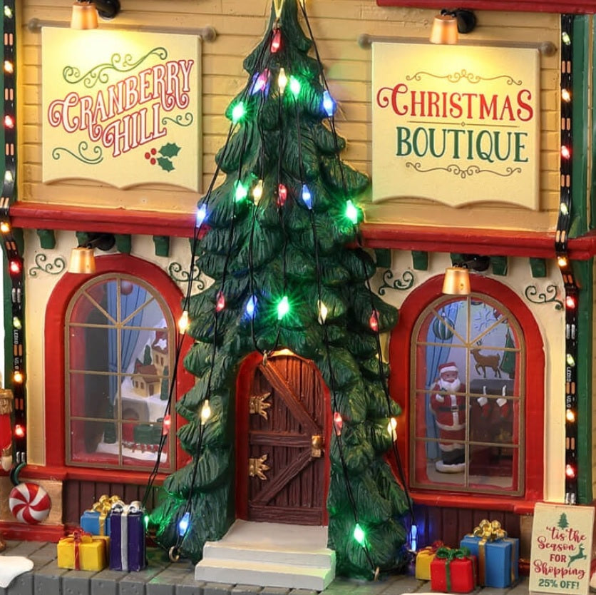 Resin lighted building for a Lemax display. Designed to look like a Christmas store. There are decorative ornaments on the roof, a large decorated Christmas tree over the front door, a nutcracker, presents and a snowman on the sidewalk.