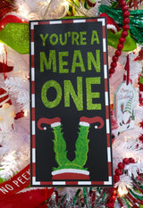 Hanging sign decoration for Christmas. The border is red and white striped. The background is black and reads "YOU'RE A MEAN ONE" and features Grinch legs with red elf shoes. The text is decorated with green glitter.
