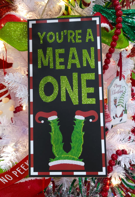Hanging sign decoration for Christmas. The border is red and white striped. The background is black and reads "YOU'RE A MEAN ONE" and features Grinch legs with red elf shoes. The text is decorated with green glitter.
