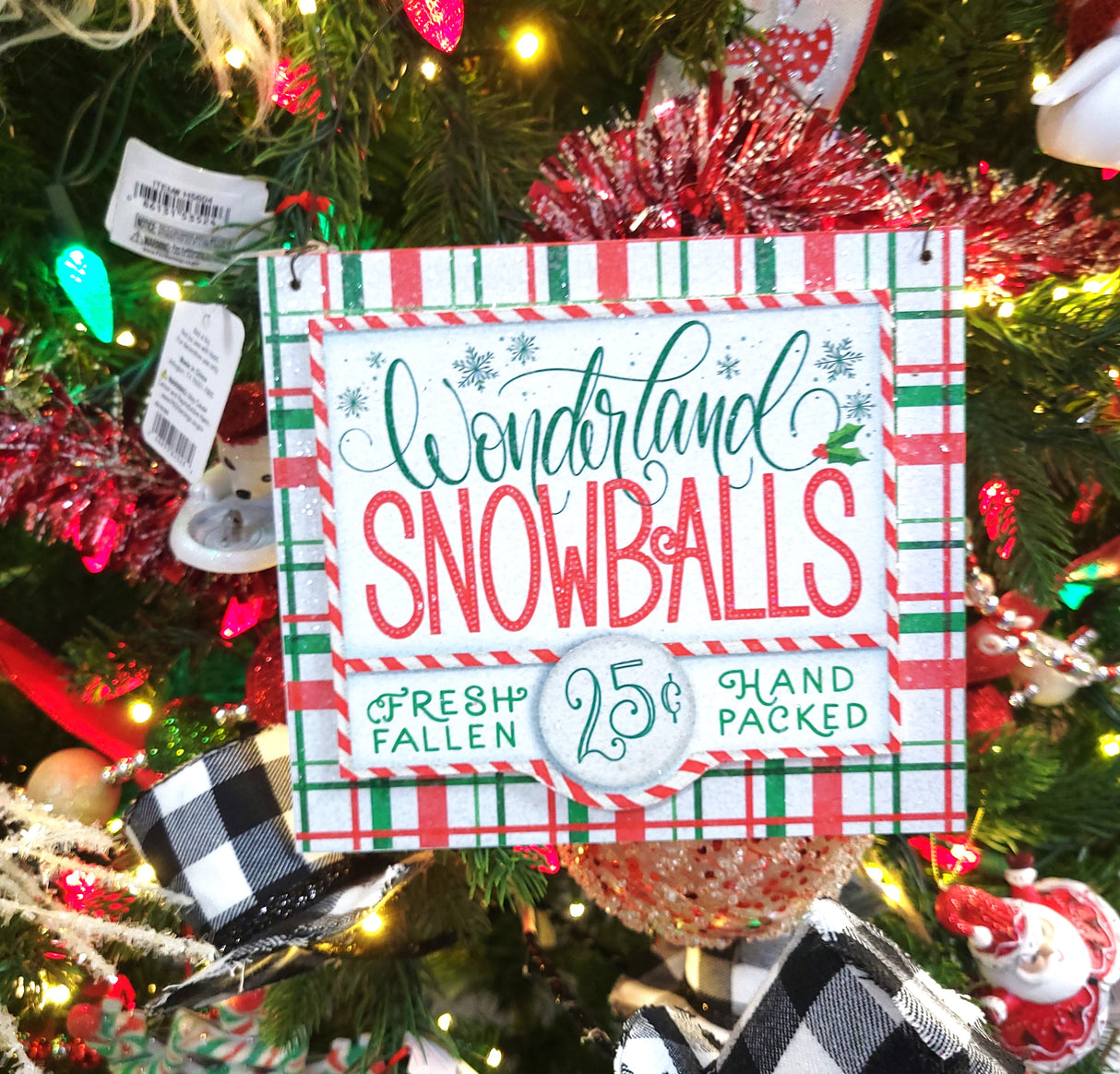 Sign hanging on a tree with other ornaments and tinsel. Sign reads "Wonderland Snowballs. Fresh Fallen. 25¢. Hand Packed."