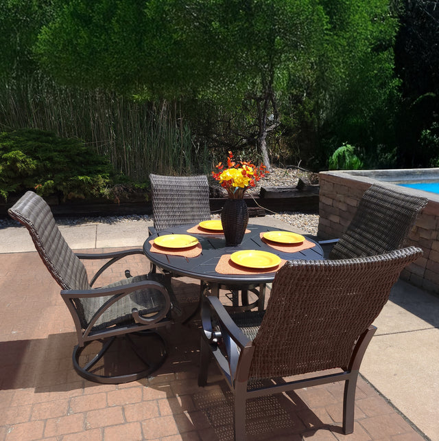 This image shows a brown wicker patio dining set with a round brown table in the center. There are four chairs. On the table, there are four yellow plates on four orange mats. In the center of the plates, there's a vase with yellow and red flowers inside.