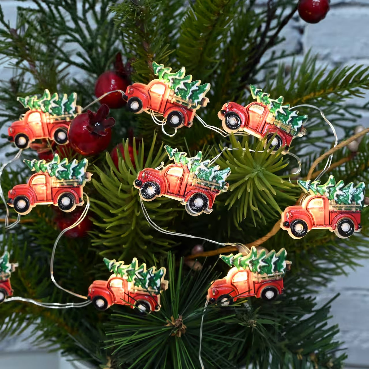 This is a lifestyle image showing the truck lights strung up on a tree. The lights consist of red pick up trucks carrying a tree covered in snow.