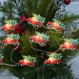 This is a lifestyle image showing the truck lights strung up on a tree. The lights consist of red pick up trucks carrying a tree covered in snow.