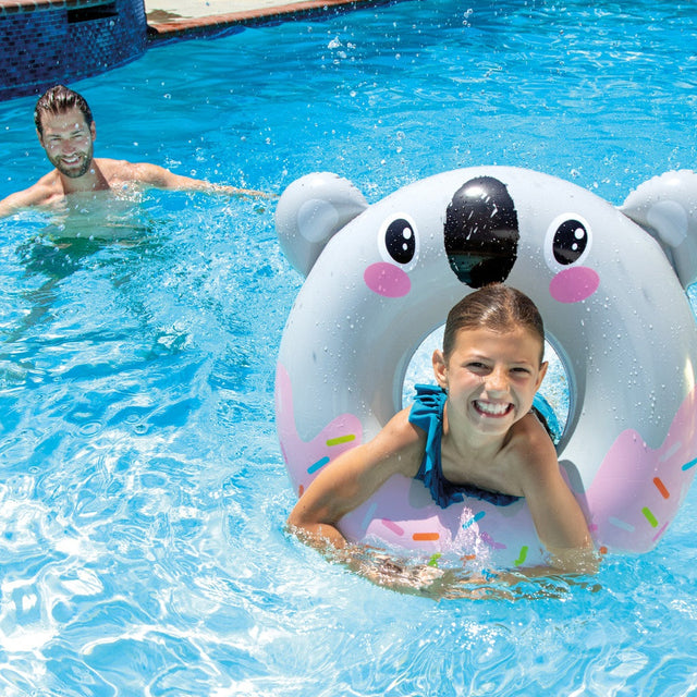 This is a lifestyle image showing a young girl inside of the koala inner tube in a pool while her dad looks on.