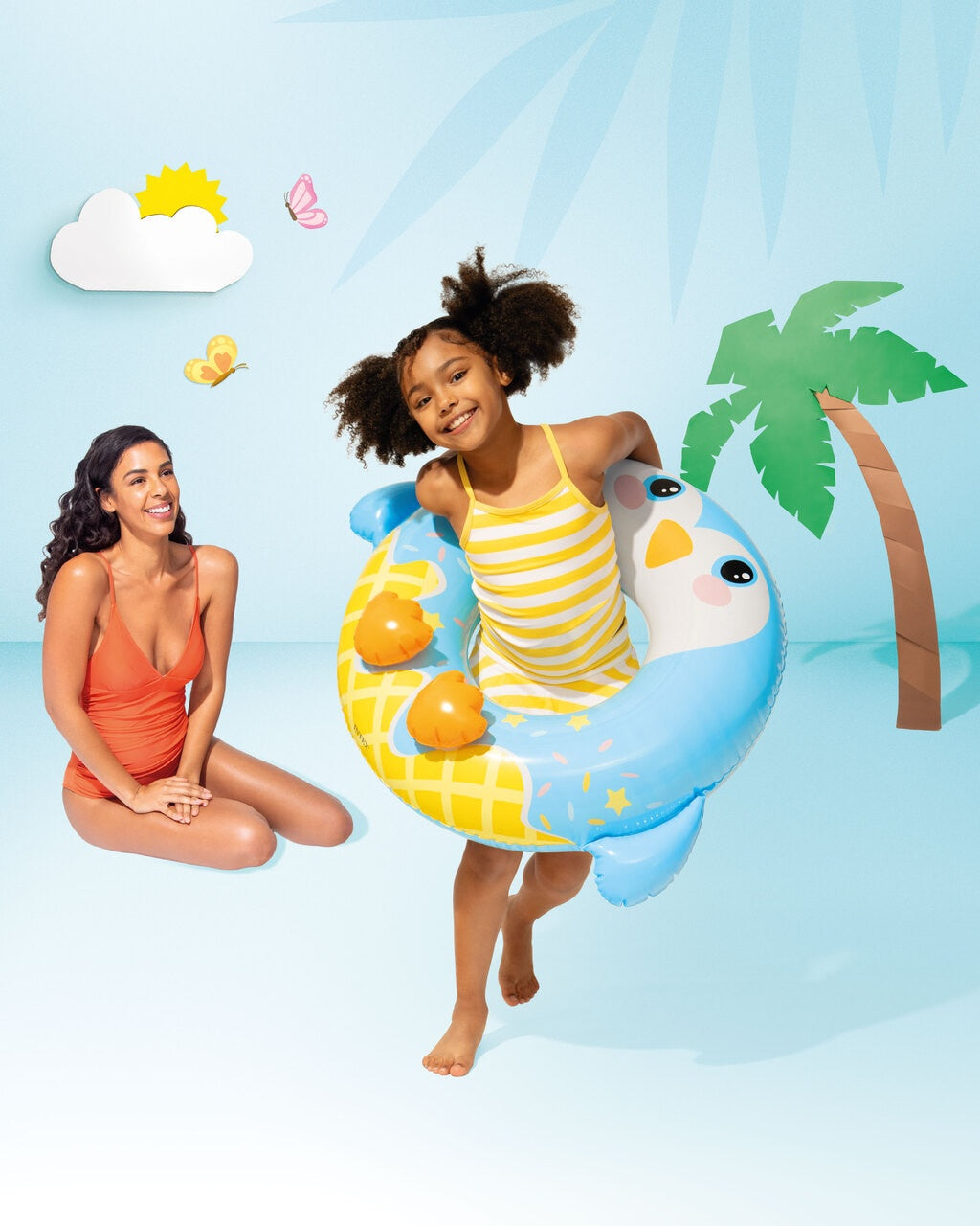 This is a lifestyle image showing a young girl inside the penguin inner tube while her mom looks on. The backdrop is blue with cartoon-ish looking details. The sun, cloud, butterflies and palm tree all look like they're made from cardboard cutouts.