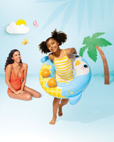 This is a lifestyle image showing a young girl inside the penguin inner tube while her mom looks on. The backdrop is blue with cartoon-ish looking details. The sun, cloud, butterflies and palm tree all look like they're made from cardboard cutouts.