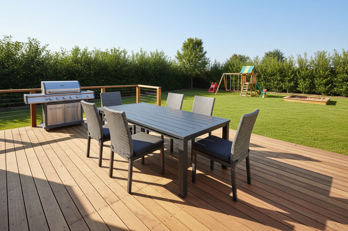 This is a lifestyle image showing this patio dining set on a wooden deck in a grassy backyard. On the deck is also a silver grill. In the backyard is also a playset and a sandbox.