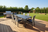 This is a lifestyle image showing this patio dining set on a wooden deck in a grassy backyard. On the deck is also a silver grill. In the backyard is also a playset and a sandbox.