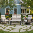 This is a lifestyle image showing this cushioned patio furniture set on a stone patio on a stone patio in the backyard of a light blue house.