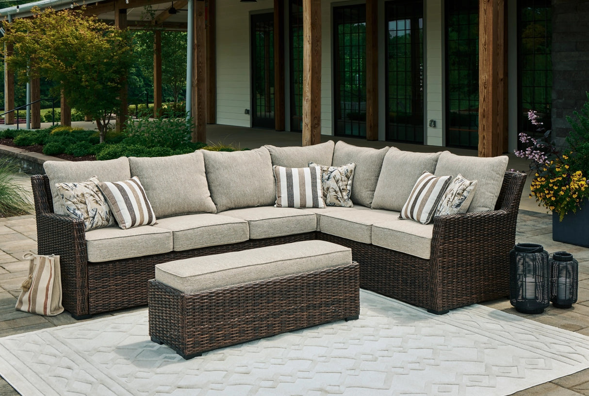 This is a lifestyle image showing the sectional sofa and the cushioned bench on a stone patio next to a house.