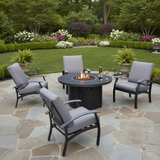 This is a lifestyle image showing four cushioned patio chairs around a circular fire pit table on a stone patio in a grassy backyard.