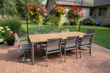 This is a lifestyle image showing this patio dining set on a brick patio in a grassy backyard. There are hanging planters of flowers and potted plants.