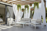 This is a lifestyle image showing the patio furniture set on a concrete patio underneath a pergola decorated with hanging plants and white, breezy curtains.