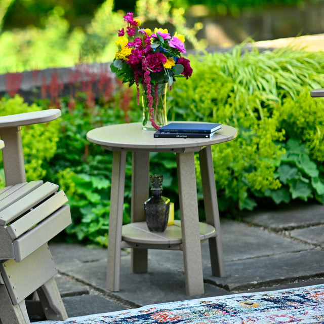 This is the Coastal Gray colored end table next to a matching chair. On top of the table is a vase of flowers and two books. On the shelf underneath the tabletop, there's a glass bottle.