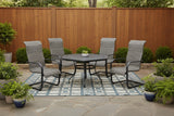 This is a lifestyle image showing these spring wicker chairs and a hexagon dining table on an outdoor rug on a stone patio in a backyard. There are lots of potted plants and a tall, wooden fence.