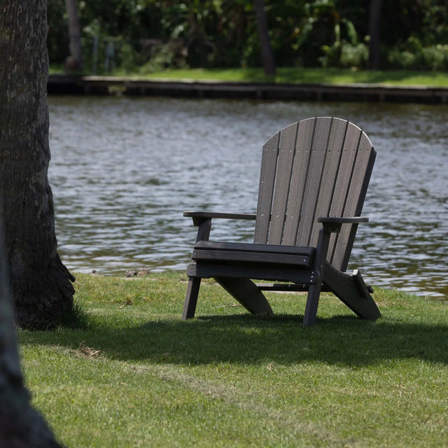 This is the Coastal Gray colored Folding Adirondack chair on a grassy patch under some trees next to a body of water.