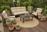 This is a lifestyle image showing this patio furniture set on a wooden porch surrounded by bushes and potted cacti plants.