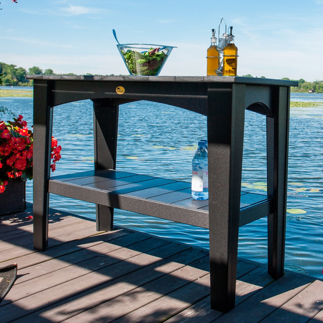 This is a lifestyle image showing the Black colored Island Buffet Table on a wooden dock overlooking a body of water. On top of the table is a bowl of salad and two containers of salad dressing.