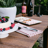 This is an up close lifestyle image showing the Antique Mahogany colored Island Buffet Table. On top of the table are a cake with berries on it, a vase, a serving spoon, a serving of small cookies and two bottles of wine.