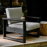 This is a lifestyle image showing the deep seating chair with a black frame and a spectrum dove colored cushion. The chair is on a wooden porch. There's a small cup of coffee on the armrest.