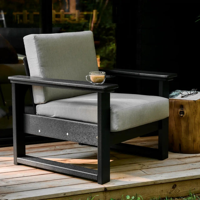 This is a lifestyle image showing the deep seating chair with a black frame and a spectrum dove colored cushion. The chair is on a wooden porch. There's a small cup of coffee on the armrest.