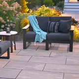 This is a lifestyle image showing the deep seating loveseat on a stone patio in a backyard with lots of plants. Off to the side of the image, you can see a small, circular table and a deep seating chair. The frames are black and the cushions are spectrum carbon.