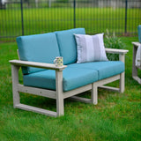 This is a lifestyle image showing the deep seating loveseat in a grassy yard. There's a white and tan striped throw pillow on the loveseat and a mug on the armrest. The frames of the loveseat are dove gray and the cushions are cast breeze.
