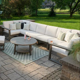 This is a lifestyle image showing the deep seating sectional set on a brick patio in a grassy backyard on a sunny day. The frames are coastal gray and the cushions are canvas.