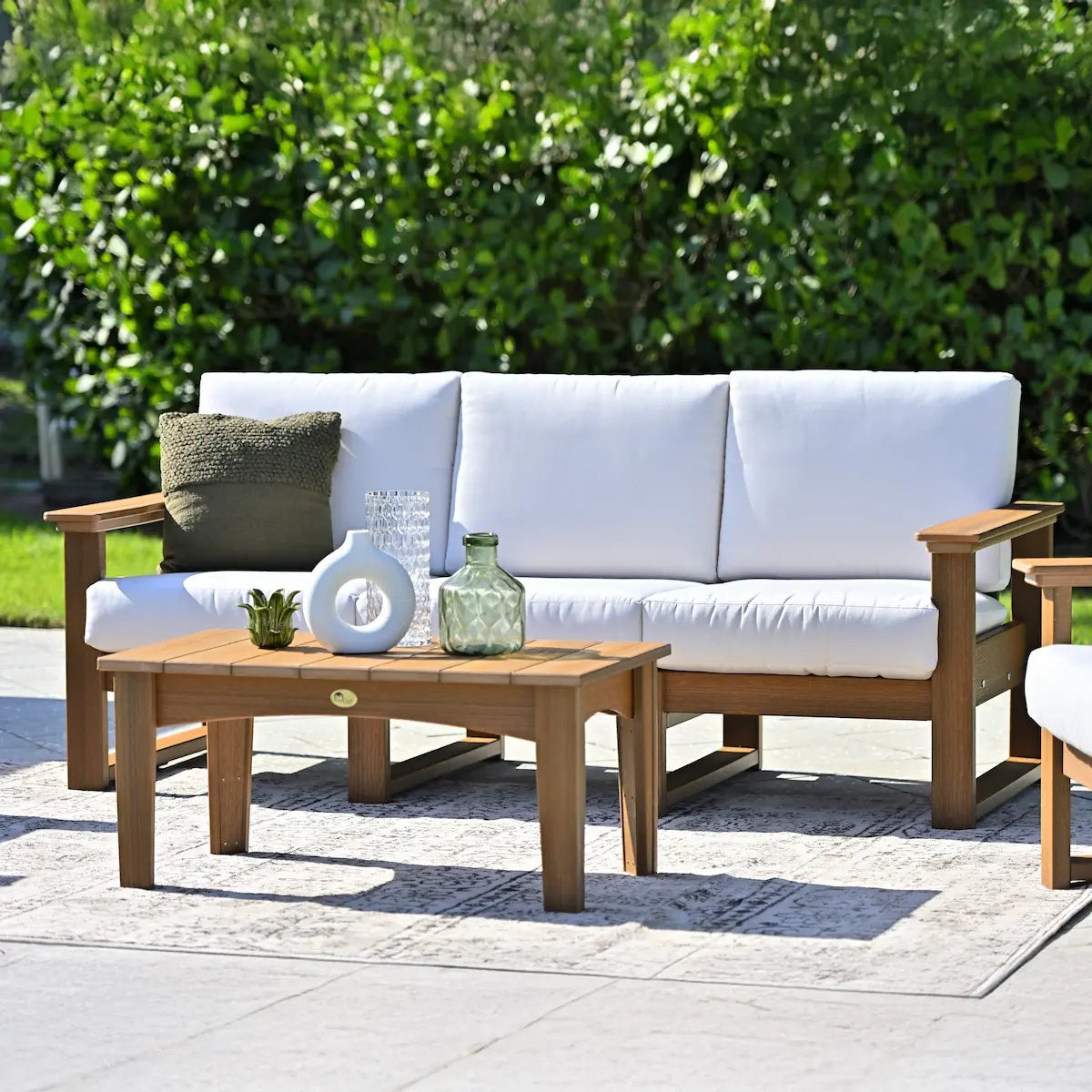 This is a lifestyle image showing the deep seating sofa on an outdoor carpet on a stone patio in a grassy backyard on a sunny day. The frames are antique mahogany and the cushions are canvas.