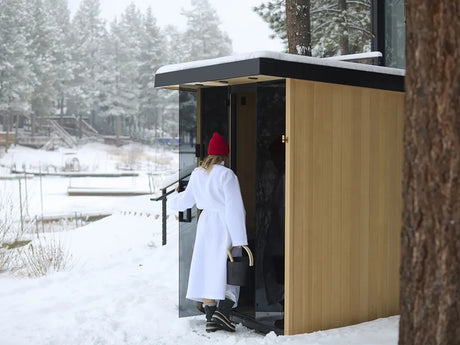 This is a lifestyle image showing a woman opening the door to her outdoor sauna on a snowy day. She's wearing a red winter hat, a white robe and winter boots.
