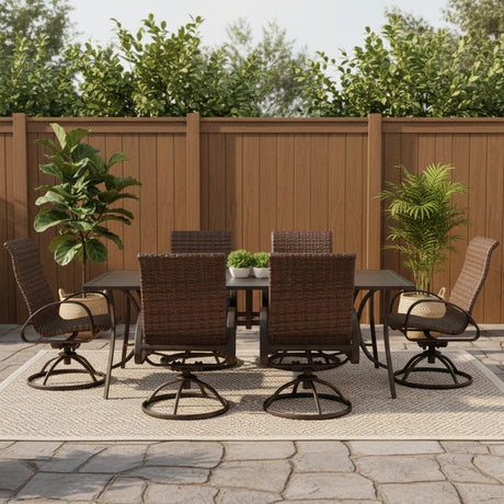 This is a lifestyle image showing this wicker patio dining set on an outdoor rug on a stone patio. There are potted plants and a tall, brown fence.